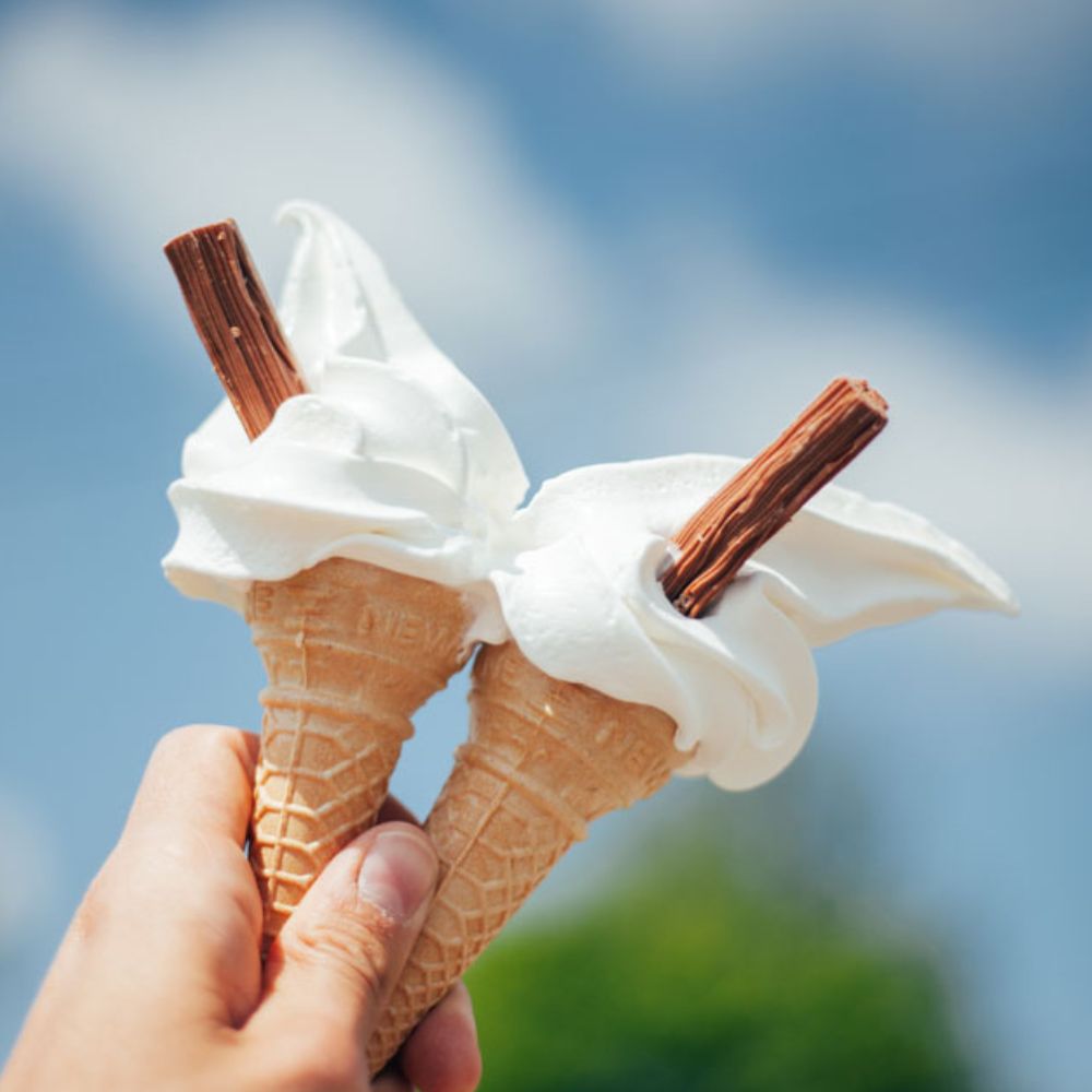 Two ice cream cones with white whipped cream and flakes held against a blue sky.